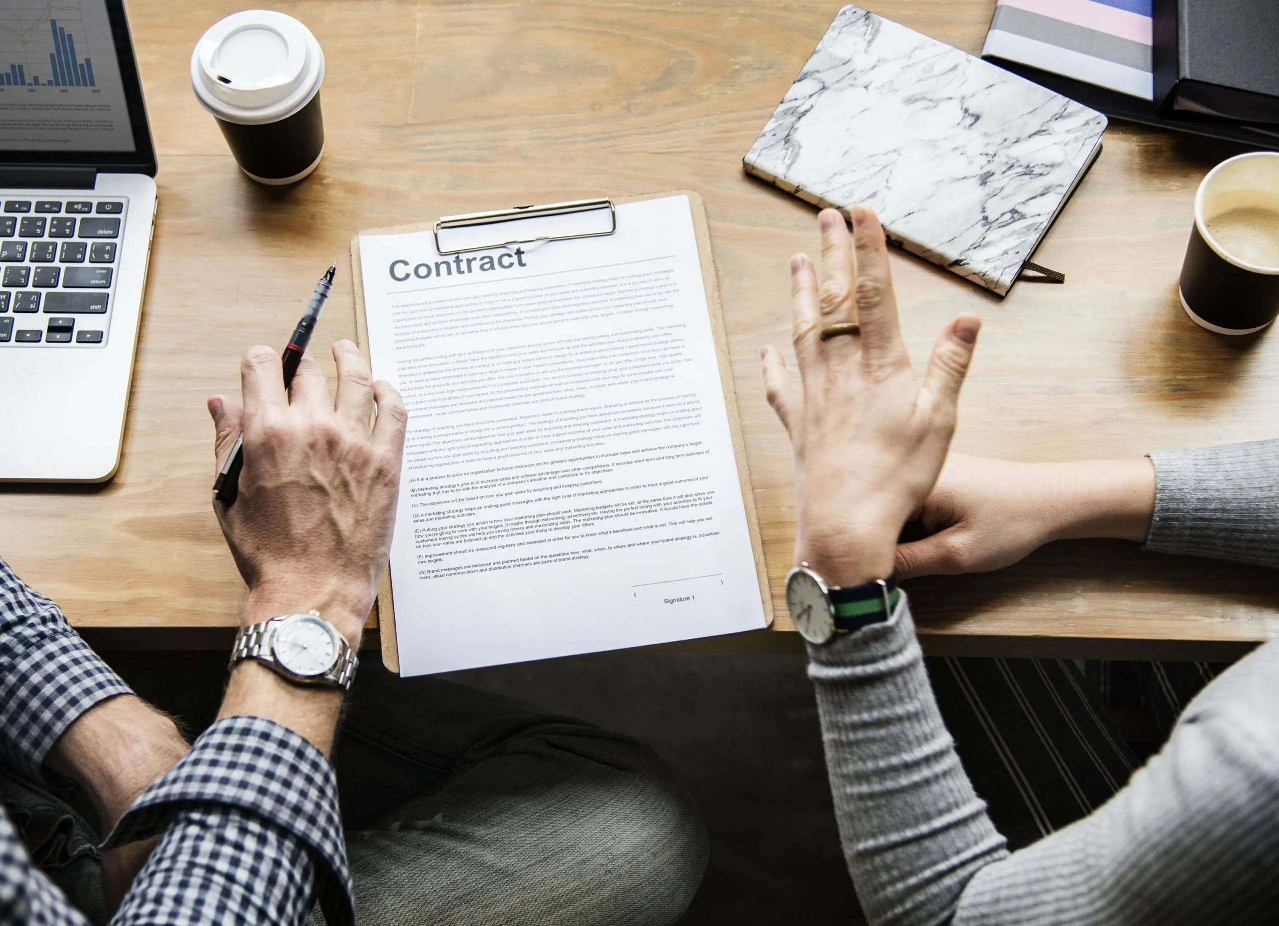 This overhead photograph captures two people engaged in a professional discussion centered around a contract on a wooden desk. The image features several key elements: The Contract: A document titled 
